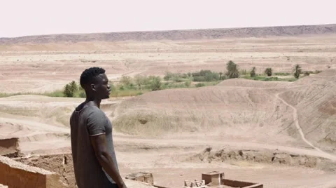 Black guy on a high peek of sand houses and looks around at the desert Stock Footage 67557962