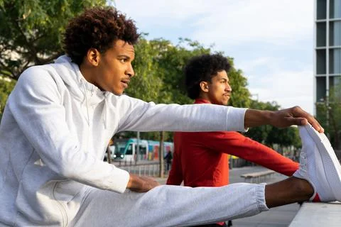 Black guy stretching hamstrings near African American friend on street Stock Photos
