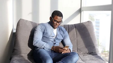 Black guy uses a smartphone sitting on couch Stock Footage 77242824