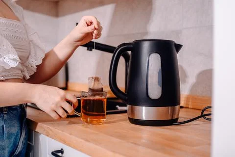 A black kettle is on the kitchen table. A woman's hand holds a teapot and mak Stock Photos