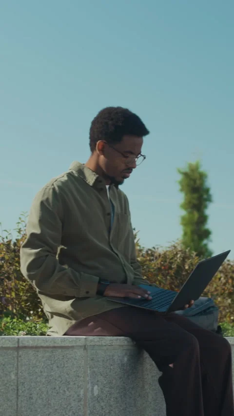 Black Male Programmer Sitting on Low Concrete Wall in Park and Working on Laptop Stock Footage 303629429