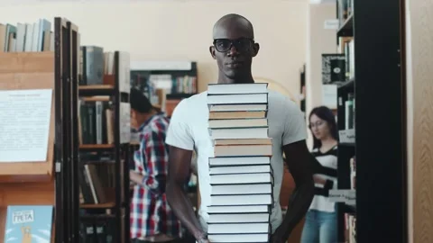 Black male student stands with a stack of books in library against the Stock Footage 234004102