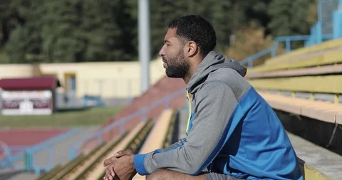 A black man with a beard and a tracksuit sits on a stadium in the fall. Vídeo Stock 101669952