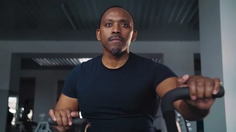 Black man doing cardio workout sitting on bicycle machine in crossfit gym Stock Footage 225713081