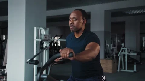 Black man doing cardio workout sitting on bicycle machine in crossfit gym Stock Footage 225713357