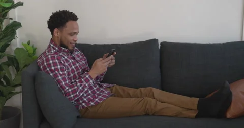 Black man at home using smartphone texting sitting on a couch Stock Footage 169427231