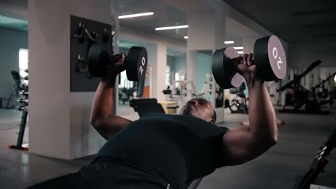 Black man lying on bench and doing dumbbell press exercise while working out in Stock Footage 225716912