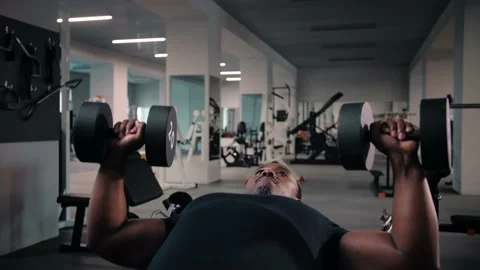 Black man lying on bench and doing dumbbell press exercise while working out in Stock Footage 225717032