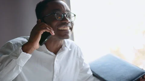 Black man in office talks on smartphone with humor and smiles while explaining 스톡 동영상 335755041