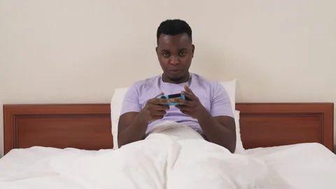 Black man playing console in bed. white woman's legs appear in front of Stock Footage 136586821
