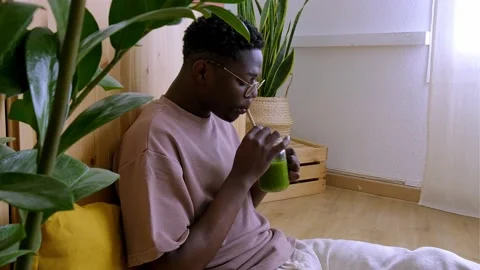 Black man relaxing at home drinking green juice sitting on the floor. 4k video. Stock Footage 234656317