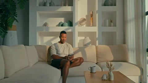 Black Man Relaxing on Sofa scrolling his phone, Sipping Drink, then Enjoying Stockbeeldmateriaal 331111253