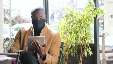 Black man sitting in a coffee table using tablet wearing face mask. Stock Footage 157323024
