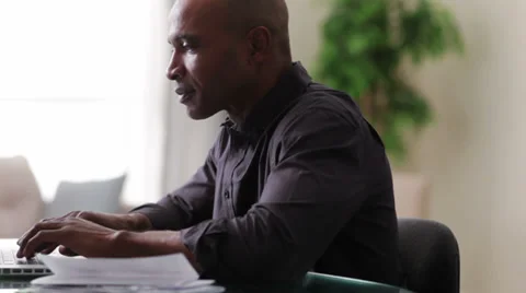 Black man sitting at table using laptop and holding paperwork Видео 36188850