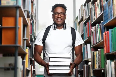 Black man stands in the library with a stack of books in his hands and smiles. Stock Photos