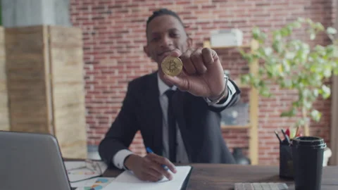 Black man in a strict suit smiling shows a bitcoin coin with hand and begins to Stock Footage 158077655