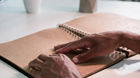 Black man studying a book for people with vision problems sitting at table at Stock Footage 254709093