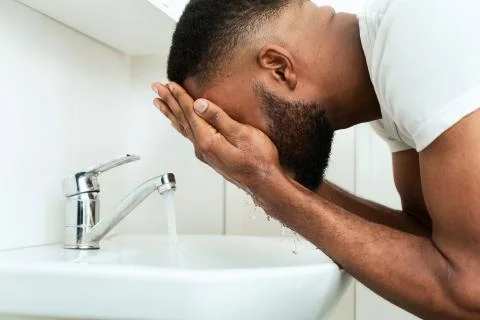Black man washing his face, splashing water at sink in bathroom Stock Photos