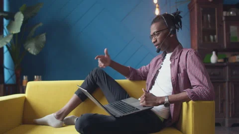 Black man wearing a headset is sitting in relaxed position the couch with laptop Stock Footage 155550873