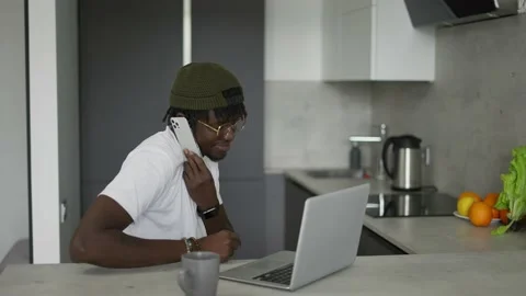 Black man working at home in kitchen talking on smartphone and using laptop Stock Footage 198700969