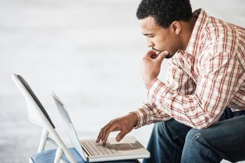 Black man working on a lap top computer and usiing a chair as a table top. Foto stock
