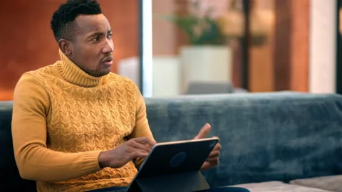 Black man working using a tablet while talking with other people, sitting on a Stock Footage 235209038