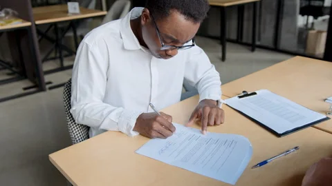 A black man writes his name on a contract in an office. He is focused while Stock Footage 335733632