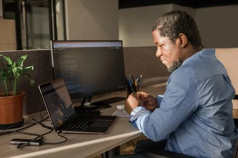 Black Man Writing Lines of Code On Desktop PC With Two Monitors and a Laptop Stock Photos