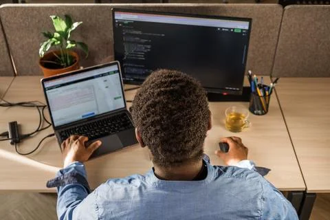 Black Man Writing Lines of Code On Desktop PC With Two Monitors and a Laptop Stock Photos