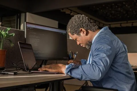 Black Man Writing Lines of Code On Desktop PC With Two Monitors and a Laptop 写真素材