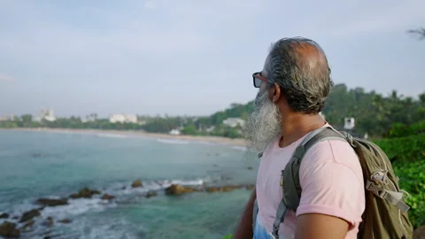 Black old man with backpack watching landscape of blue ocean with rocks at Stock Footage 256011219