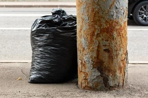Black plastic garbage bag leaning against a rusty metal pole on the city street Stock Photos