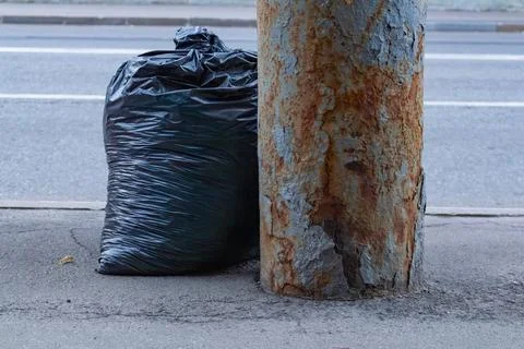 Black plastic garbage bag left on the city street next to rusty metal pole Stock Photos