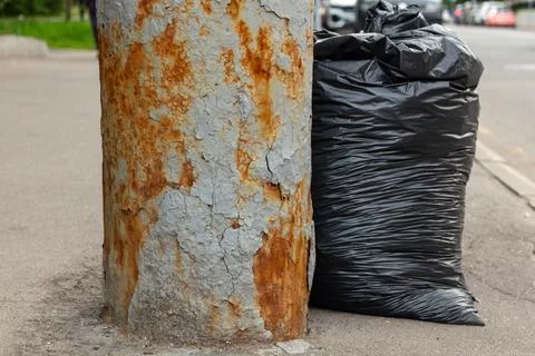Black plastic garbage bag standing next to old rusty metal pole on city street Stock Photos