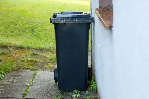 Black plastic garbage bin on wheels standing next to a house wall outdoors. Stock Photos