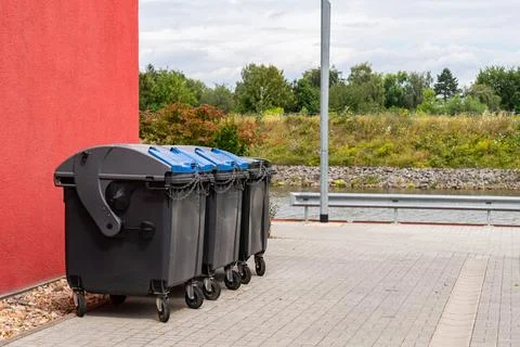 Black plastic trash cans closed on a chain stand near a large red building. Stock Photos