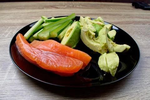 On a black plate, sliced cucumber, avocado and salted salmon. Preparations fo Stock Photos