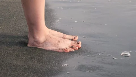 Black sand beach with gentle waves washing over woman's bare feet, creating Vidéo 301920700