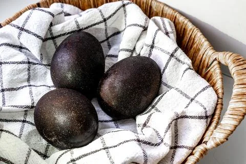 Black shiny easter eggs on a white towel in a brown basket on white background Stock Photos