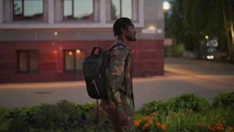 Black Student Glancing At Building Facade At Dusk, Camo Jacket And Backpack Vídeos de archivo 327229499