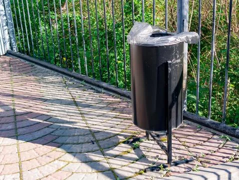 Black trash can placed on a paved pathway near a green area with a fence in Stock Photos