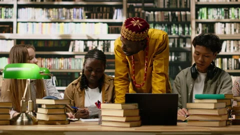 Black tutor leaning over shared study desk with laptop and open books Stock Footage 337232504