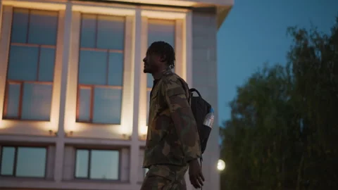 Black Walker Passing Lit Modern Building Facade At Dusk, Camo Jacket And Vídeos de archivo 329494642