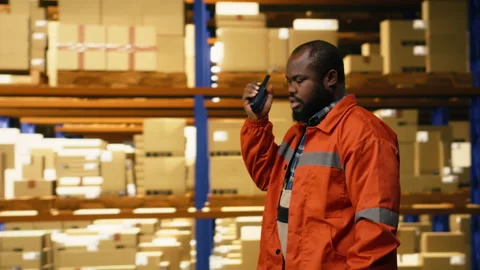 Black warehouse operator using portable walkie-talkie radio in distribution hub Stock Footage 332911824