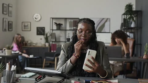 Black Woman Applying Lipstick Using Smartphone at Office Desk Video stock 345809766