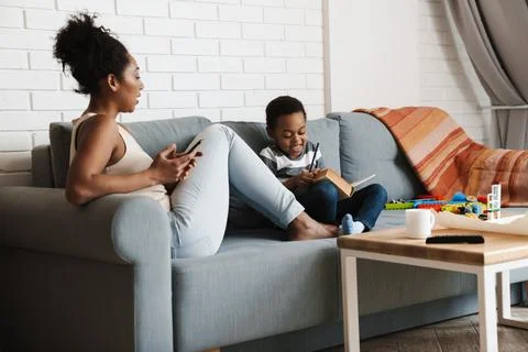 Black woman using cellphone while her son reading book at home Stock Photos
