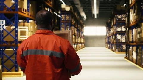 Black worker moving cargo pallets across busy warehouse floor Stock Footage 332908432