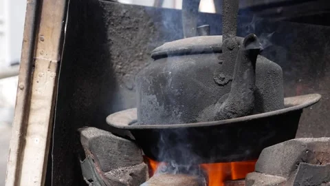 A blackened kettle heats over fire inside a clay stove while water boils. Cookin Stock Footage 338001237