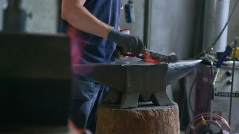 Blacksmith Brushing Hot Metal on Anvil – Glowing Forge in Background Stock Footage 313845733