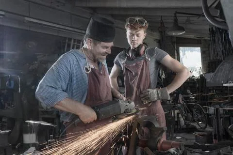 Blacksmith explaining young trainee handling of angle grinder at workshop Photos